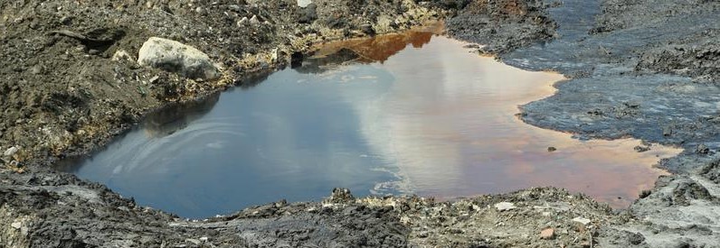 Oil-contaminated soil with a puddle reflecting the sky, surrounded by dark, polluted ground and rocks.