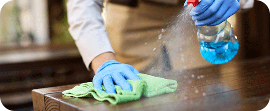 Person in blue gloves cleaning a wooden surface with a spray bottle and green cloth.